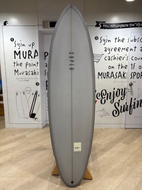 IMG_7950 - SHOP NEWS | ムラサキスポーツオンラインストア 通販 Gray surfboard on a wooden display stand inside a surf shop, with bold wall art and signage in the background.