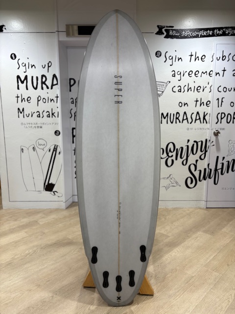 IMG_7951 - SHOP NEWS | ムラサキスポーツオンラインストア 通販 Light gray surfboard on a wooden stand in a shop, centered stringer with four fin boxes at the tail.