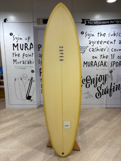 IMG_7955 - SHOP NEWS | ムラサキスポーツオンラインストア 通販 Yellow surfboard standing upright on a wooden rack indoors, with 'SUPER' along the center and surfing-themed wall art in the background.