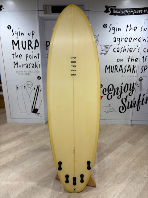 IMG_7958 1 - SHOP NEWS | ムラサキスポーツオンラインストア 通販 Yellow surfboard standing upright on a wooden display rack indoors, with black fin boxes visible at the tail and decorative wall graphics behind it.