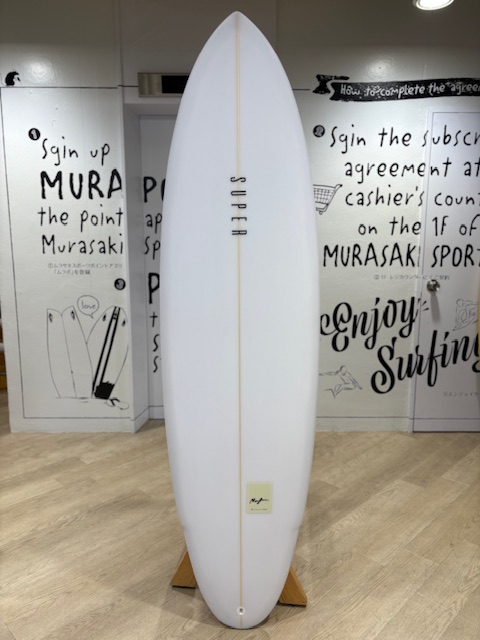 IMG_7960 - SHOP NEWS | ムラサキスポーツオンラインストア 通販 White longboard surfboard on a wooden stand inside a surf shop, with graffiti-style text on white walls in the background.
