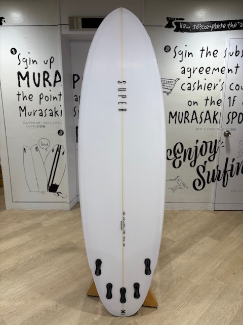IMG_7961 - SHOP NEWS | ムラサキスポーツオンラインストア 通販 White surfboard with a single stringer and four black tail fins on a wooden rack in a surf shop with graphic walls.